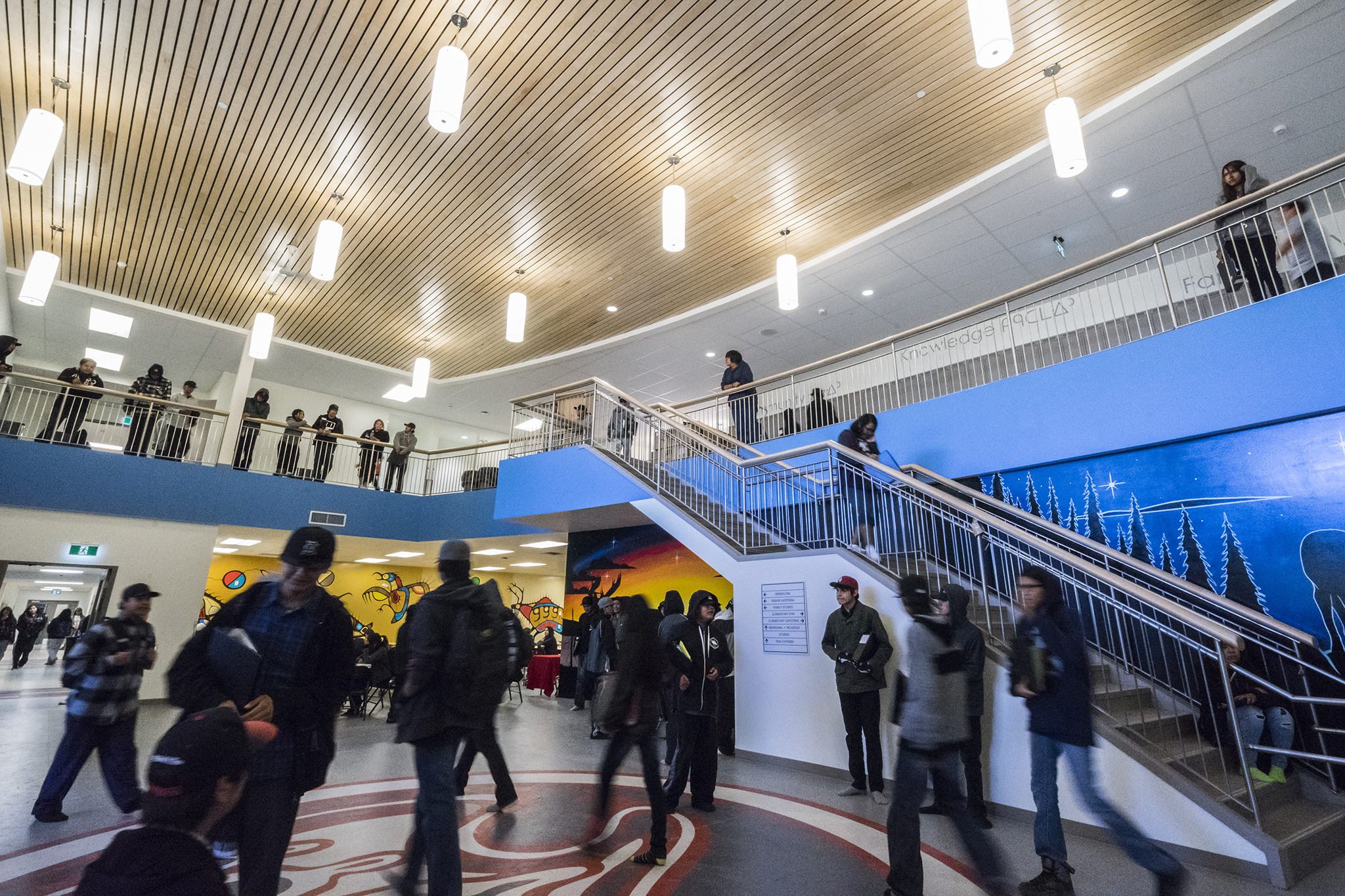 Bustling double-height foyer with a wooden ceiling treatment, wall murals, floor mural, and stairs up to a mezzanine level.