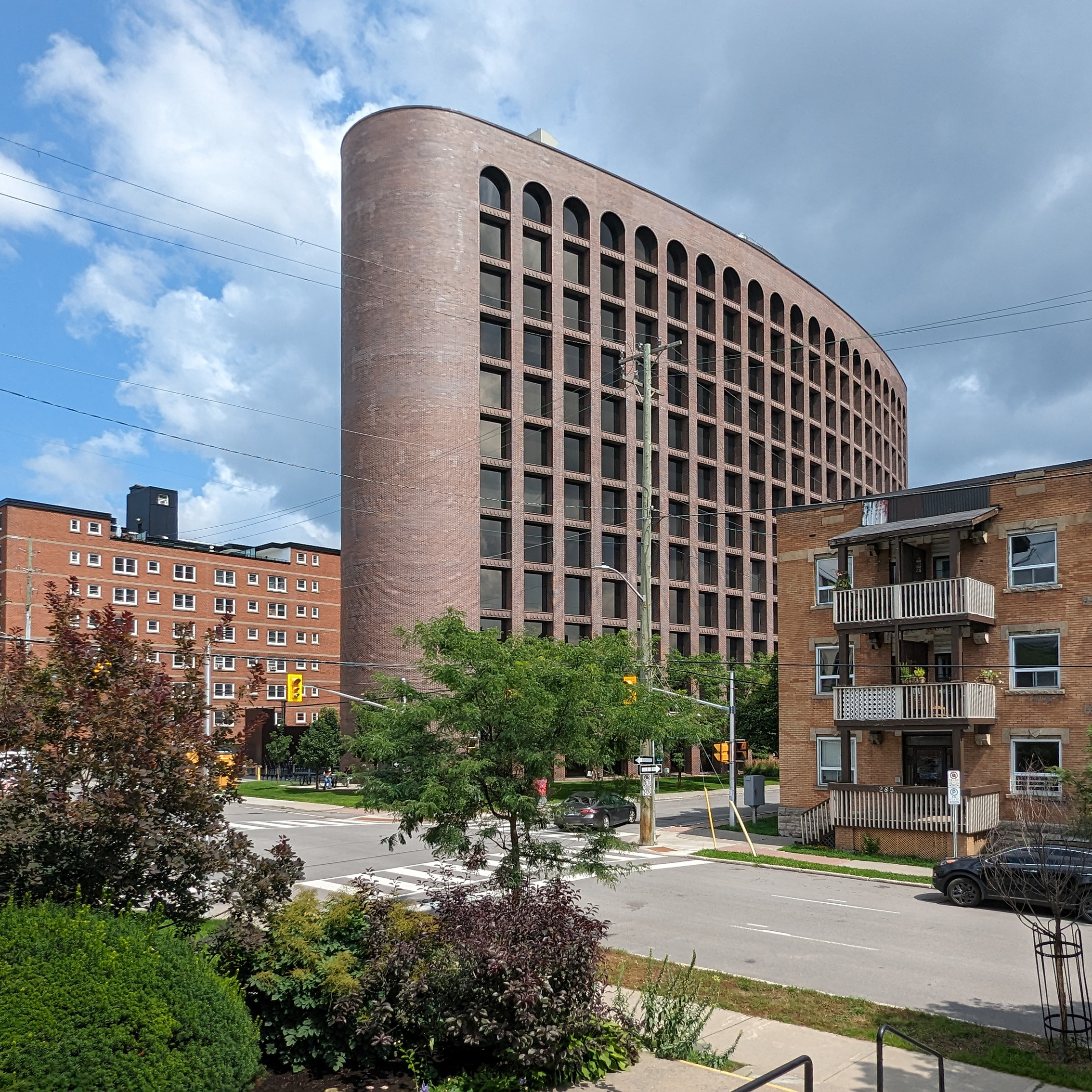 Tall and wide brown brick building in the shape of an extruded ellipse with curved walls and an orderly grid of square punch windows. The top row of windows is arched with a semicircle. Around it are 4 to 5 storey high brick buildings, rectangular in shape.